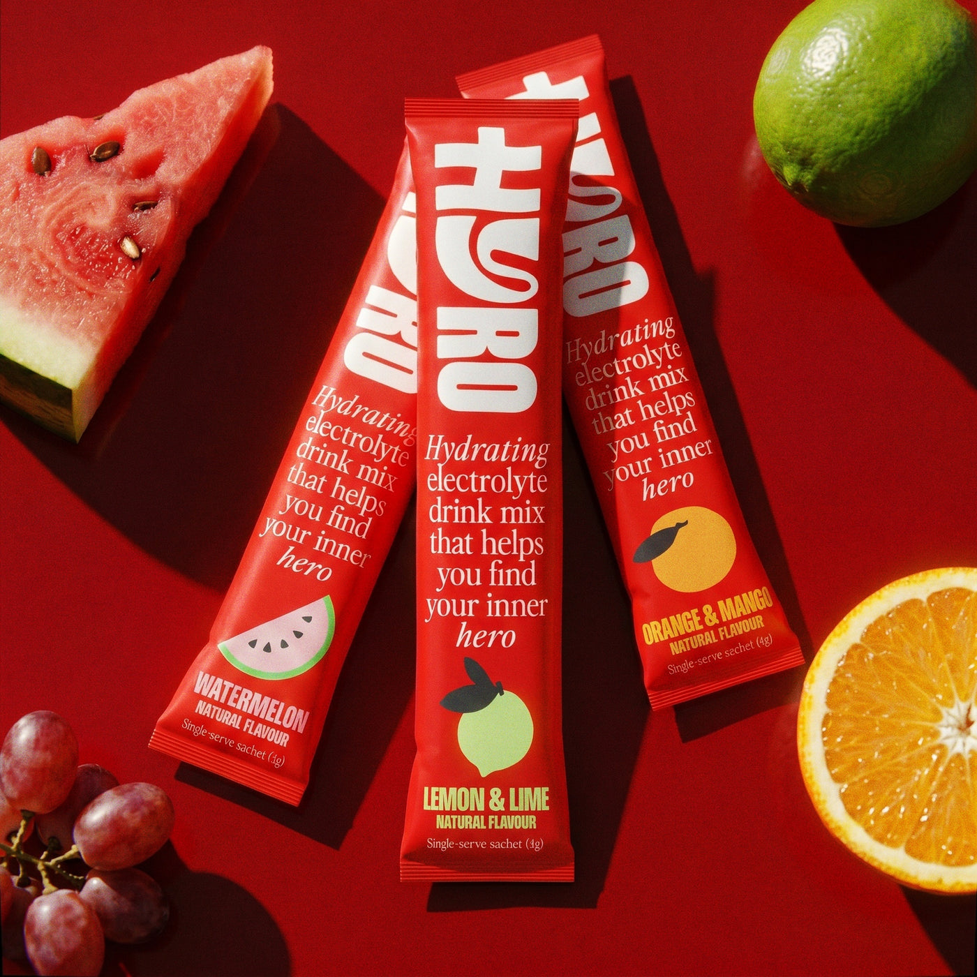 Three Hyro sticks on a red surface beside fresh watermelon, lime, orange and grapes
